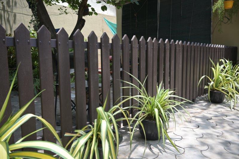 Brown wooden picket fence along a paved patio with several potted spider plants lined up beside it on a sunny day.